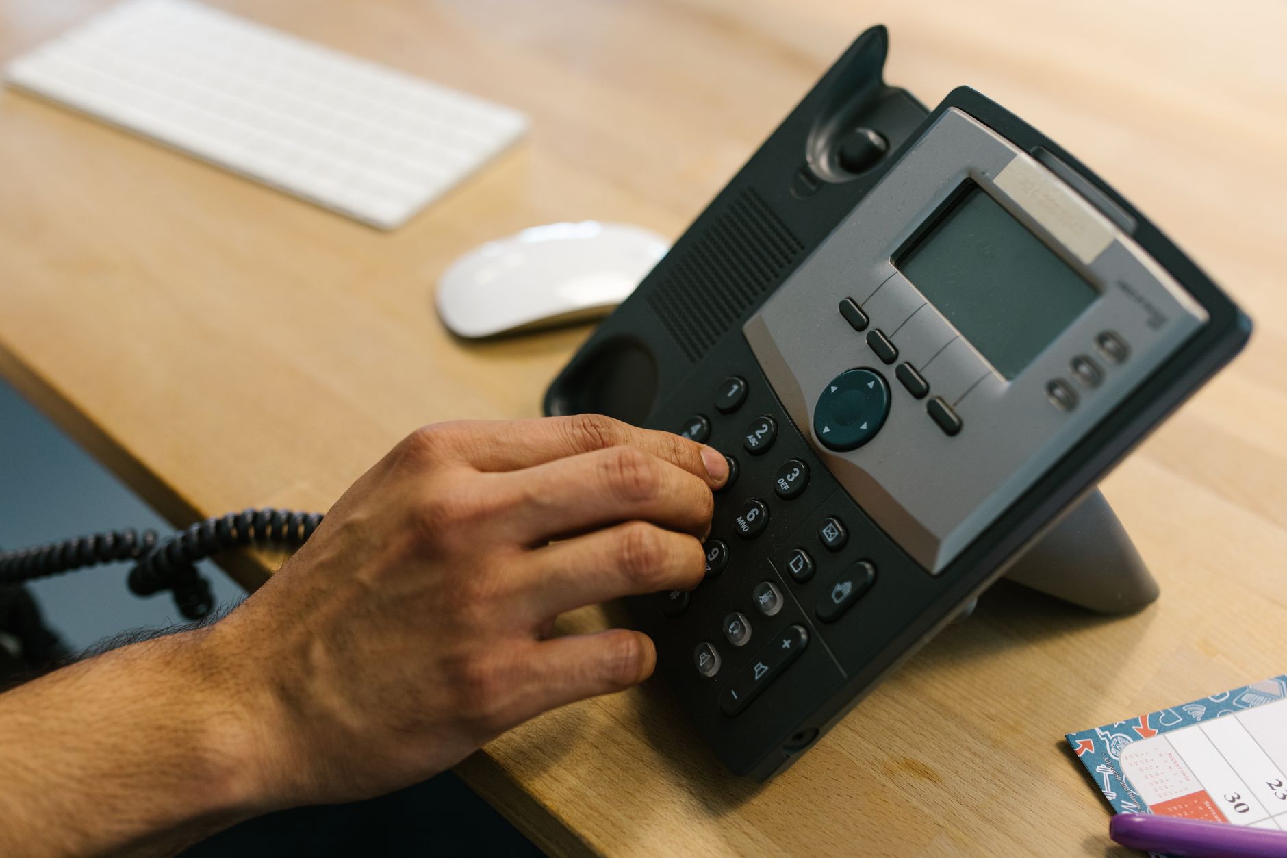main composant un numéro sur un téléphone de bureau posé sur une table en bois, à côté d'un clavier et d'une souris
