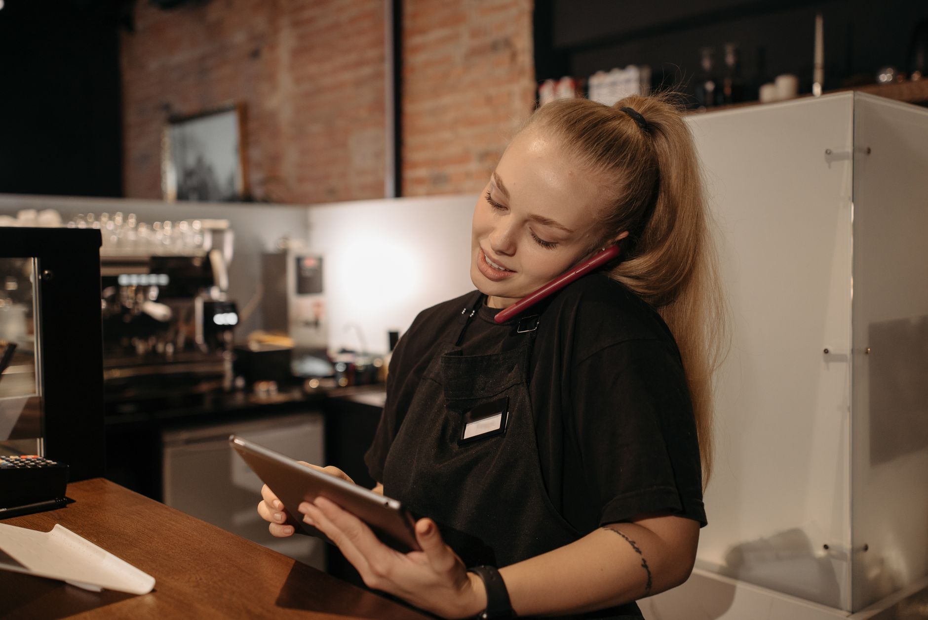 employée d'un café au comptoir, téléphone calé contre l'épaule, consultant une tablette pendant l'appel